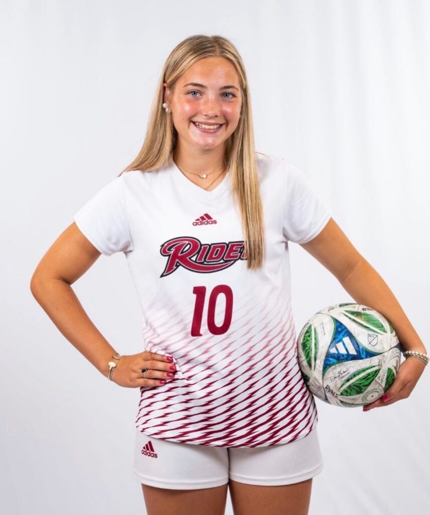 College soccer player in a white home kit holding a ball and smiling