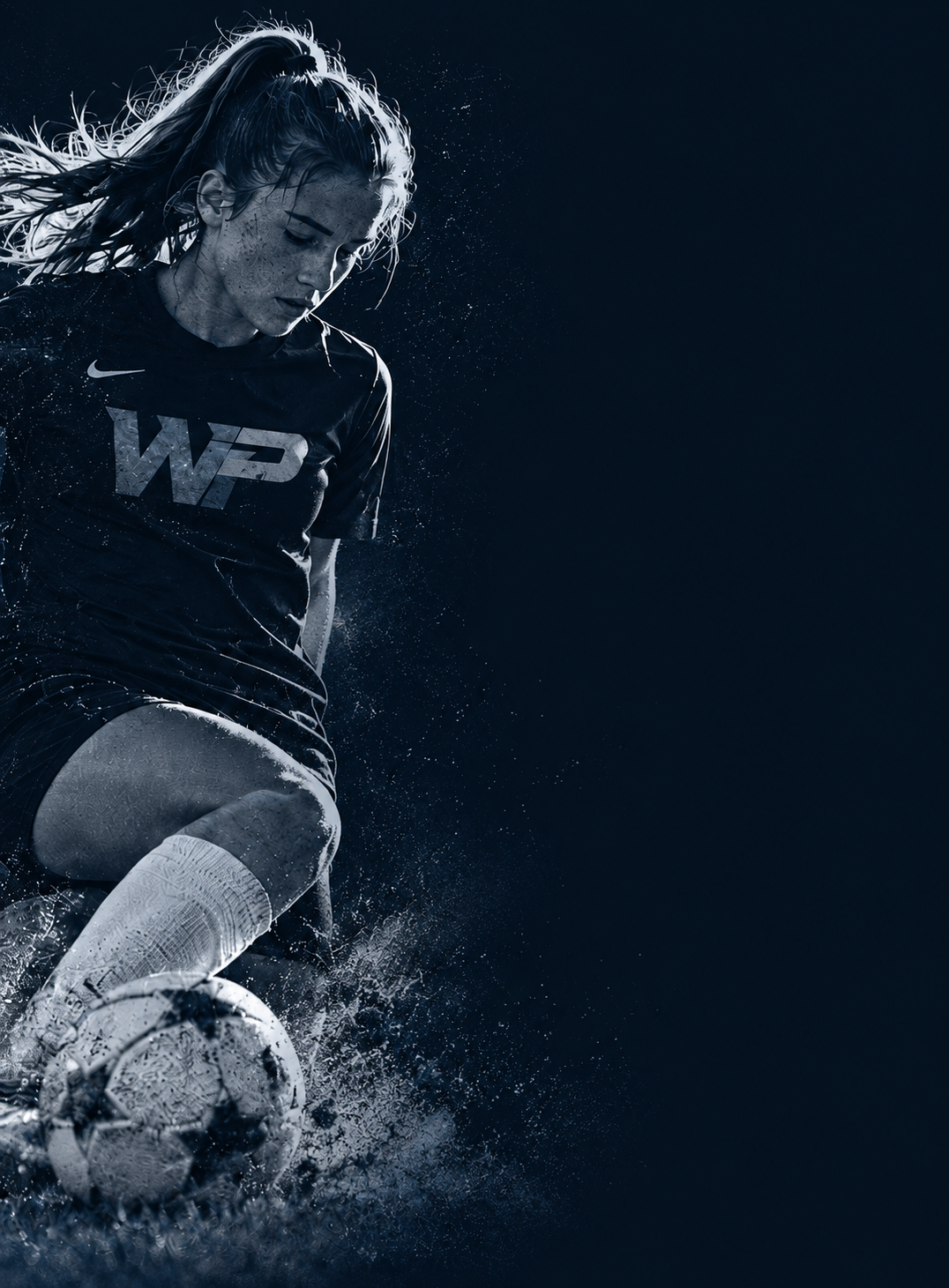 Female soccer player in a black Whole Player shirt driving through the spray with the ball